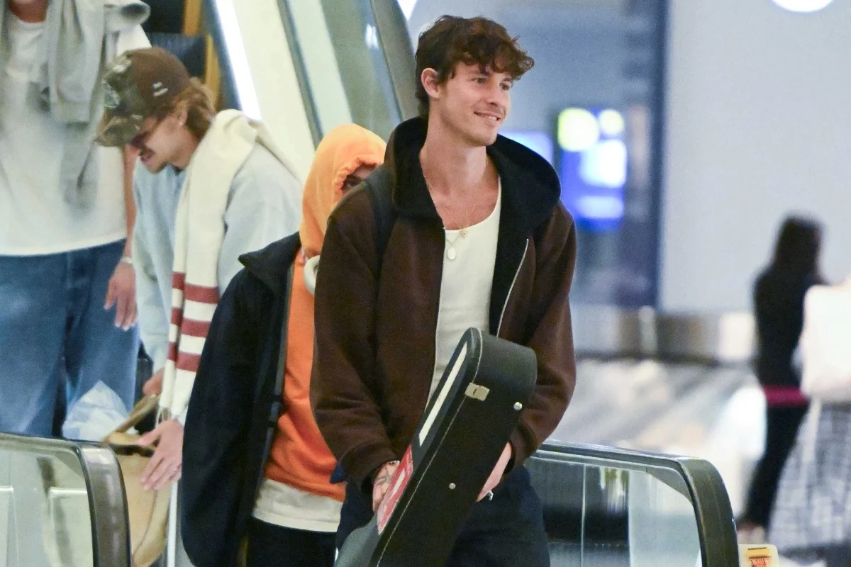 A imagem mostra o cantor Shawn Mendes descendo às escadas rolantes. Com um casaco preto e regata branca, ele carrega um vilão na mão direita e uma mochila nas costas